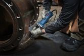A person wearing safety equipment uses a straight grinder on a metal flange.