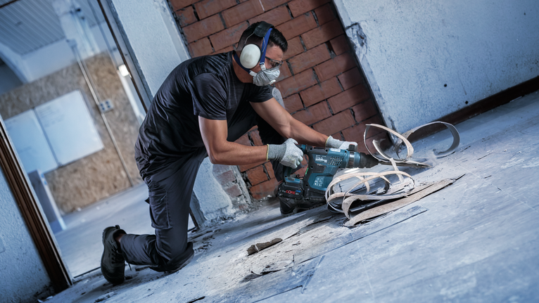 Person wearing safety equipment drills into a concrete floor near exposed wires.