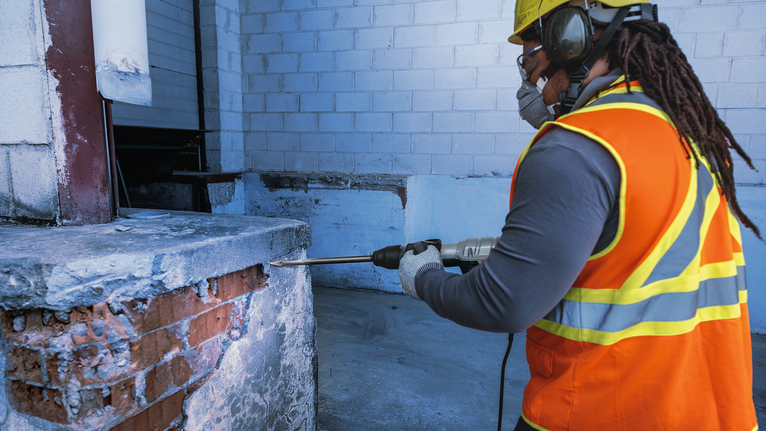 PRO SDS max-4C Pointed Chisel Set, 400 mm, 10 pcs Person wearing safety equipment drills into a concrete wall at a construction site.