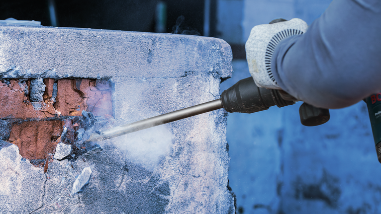 PRO SDS max-4C Pointed Chisel Set, 400 mm, 10 pcs Person wearing safety equipment uses a demolition tool to break a concrete wall.