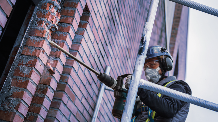 PRO SDS max-4C Pointed Chisel Set, 400 mm, 10 pcs A person wearing safety equipment drills into a brick wall on scaffolding.