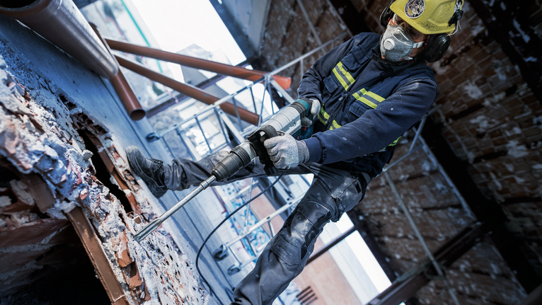 PRO SDS max-5C Flat Chisel, 25 x 400 mm A person wearing safety equipment drills into concrete at a demolition site.