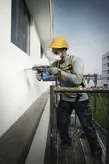 A person wearing safety equipment drills into a building wall from scaffolding.
