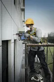 A person wearing safety equipment drills into an exterior wall while standing on scaffolding.