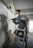 A person wearing safety equipment removes wall tiles with a rotary hammer.