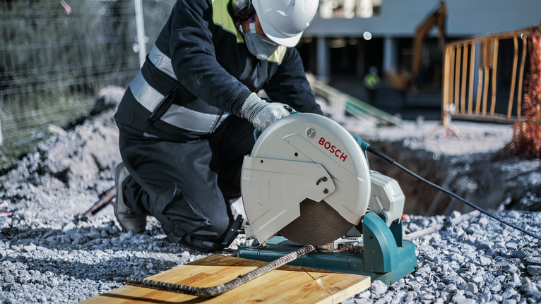 A person wearing safety equipment cuts rebar with a circular saw at a construction site.