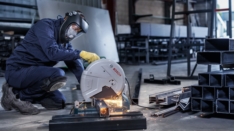 A worker wearing safety equipment cuts metal pipes indoors with a power saw.