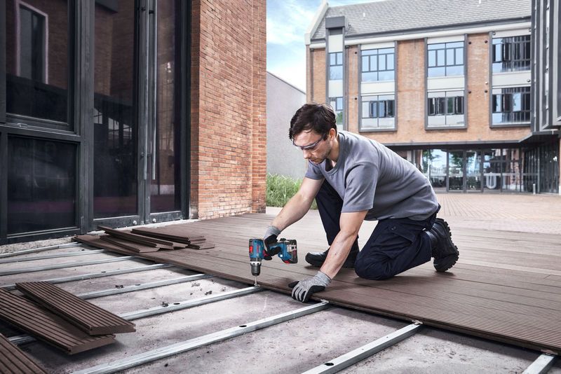 A person wearing safety equipment installs decking boards using a cordless drill.