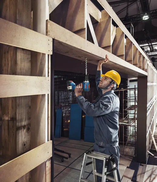 A person wearing safety equipment drills into a wooden beam while standing on a ladder.