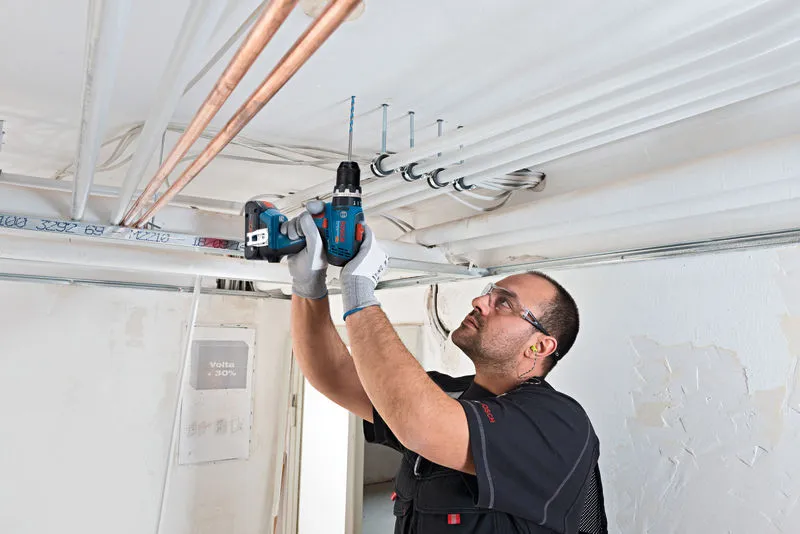 A person wearing safety equipment drills into a ceiling to install electrical conduit.