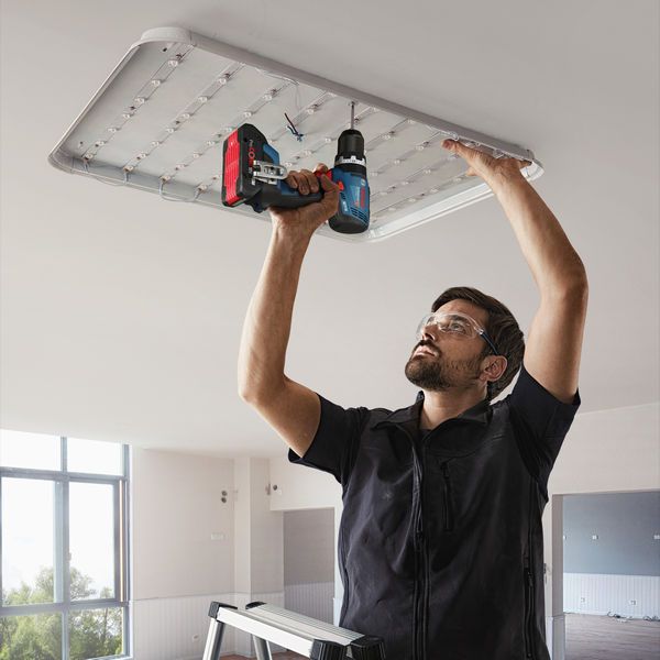 A person wearing safety equipment uses a cordless drill to install a ceiling light fixture.