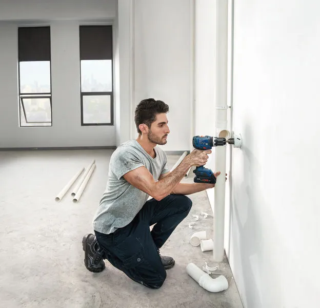 A person drills into a wall to install piping in a bright, unfinished room.