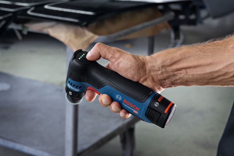 A person holds a cordless random orbit sander in a workshop setting.