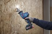 Person wearing safety equipment cuts wood panel using a cordless reciprocating saw.