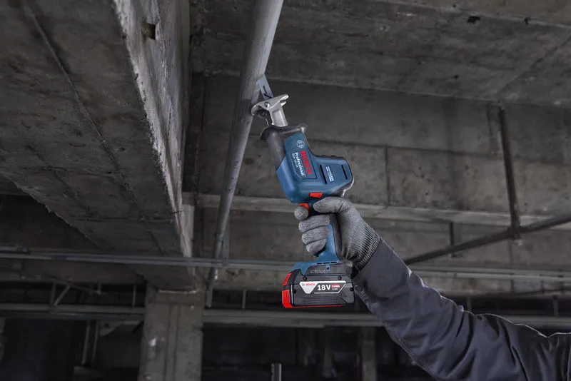 A person wearing safety equipment cuts a metal pipe overhead with a reciprocating saw.