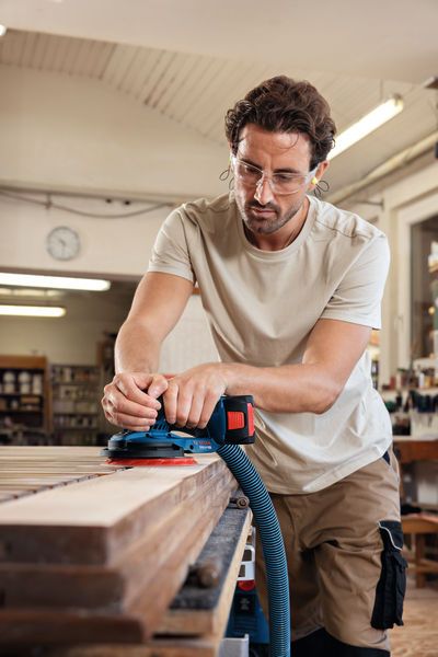 A person wearing safety equipment sands wood with a cordless random orbit sander.