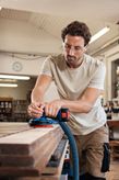 A person wearing safety equipment sands wood with a cordless random orbit sander.