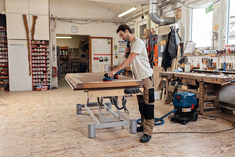 A person wearing safety equipment sands a wooden surface with a cordless random orbit sander.