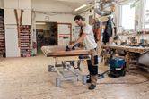 A person wearing safety equipment sands a wooden surface with a cordless random orbit sander.