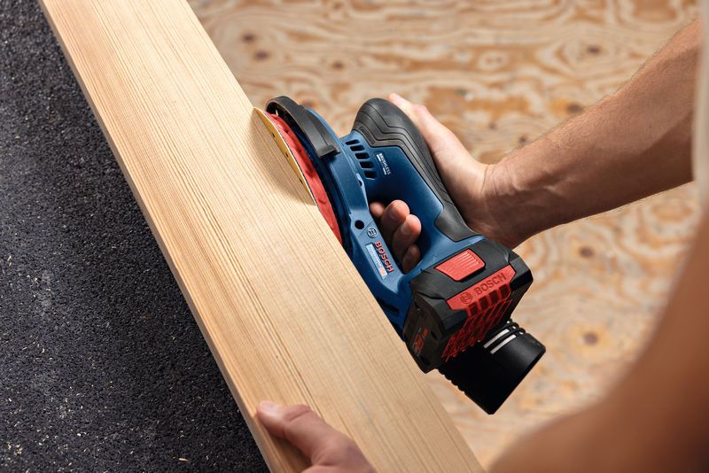 Person sanding a wooden plank using a cordless random orbit sander.