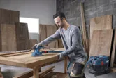 Person wearing safety equipment sands a wooden tabletop with an orbital sander.