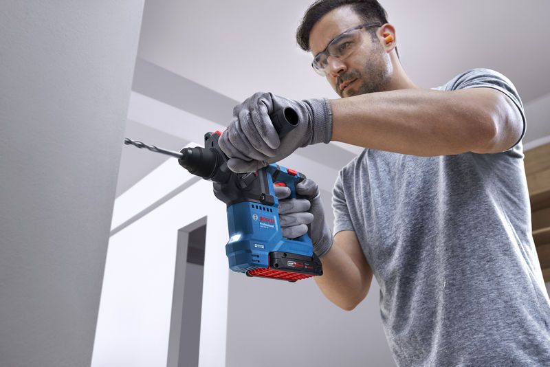 Person wearing safety equipment drills into a wall with a rotary hammer.