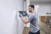 A person wearing safety equipment drills a hole into a marked white wall.