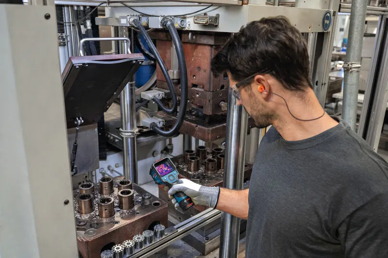 A person wearing safety equipment uses a thermo camera to inspect machinery.