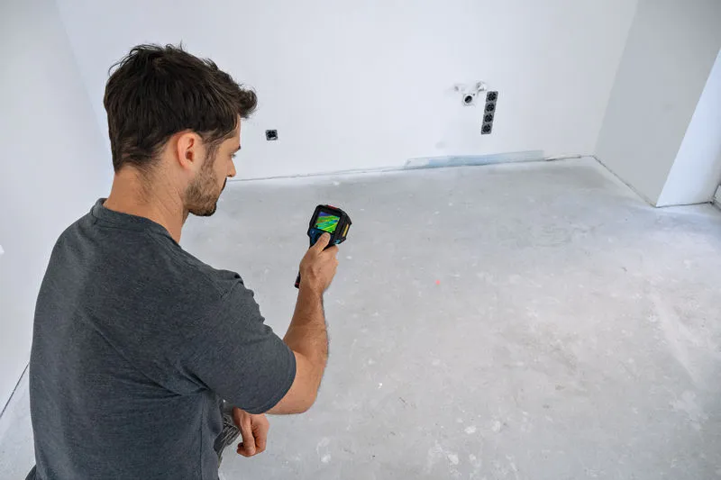 A person scans a bare room floor with a thermal camera.