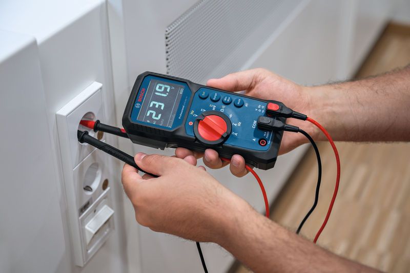 A person tests a wall electrical outlet using a digital multimeter.