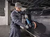 A person wearing safety equipment polishes a black car using a power tool.
