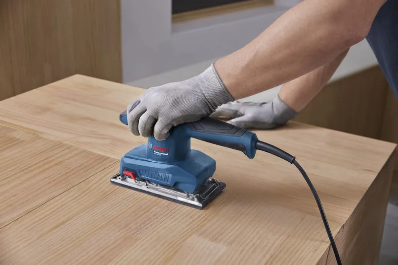 A person wearing safety equipment uses an orbital sander on a wooden surface.