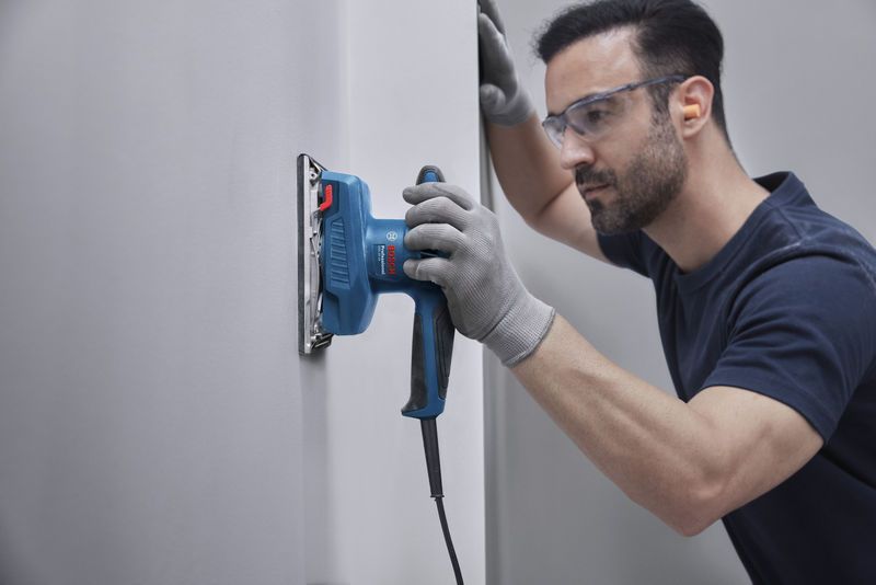 A person wearing safety equipment sands a wall with an orbital sander.