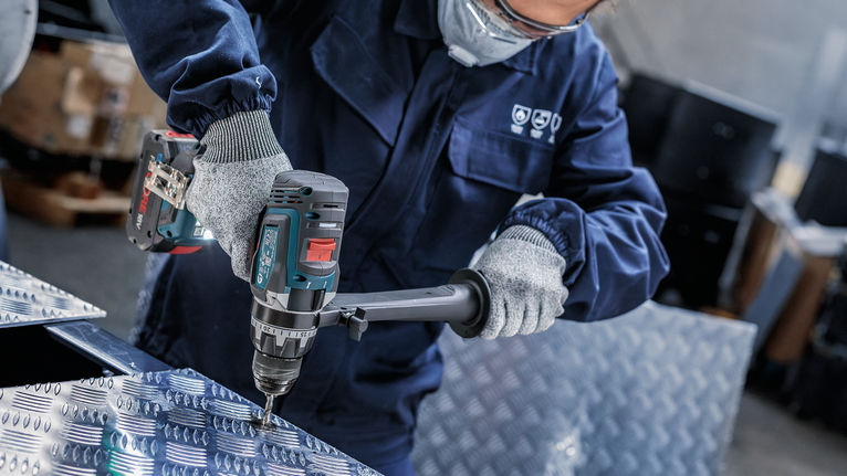 A person wearing safety equipment drills into a patterned metal sheet.