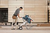 A person wearing safety equipment moves a table saw on a wheeled saw stand in a workshop.