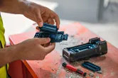 A person wearing safety equipment changes batteries in a power tool on a dusty workbench.
