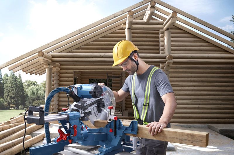 GCM 340-305 D A person wearing safety equipment cuts wood with a mitre saw at a log house construction site.