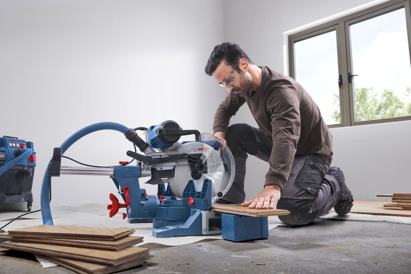 GCM 340-305 D A person wearing safety equipment cuts wooden flooring with a mitre saw in a bright room.
