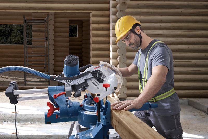 GCM 340-305 D A person wearing safety equipment cuts wood using a mitre saw at a log construction site.