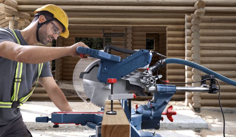 GCM 340-305 D A person wearing safety equipment operates a mitre saw to cut wood at a construction site.