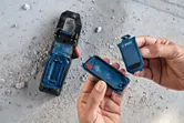 Person opens a blue battery compartment next to a tool on a dusty concrete floor.