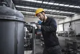 A person wearing safety equipment uses a cordless impact wrench on a large metal pipe.