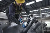A person wearing safety equipment uses a cordless impact wrench on industrial piping.