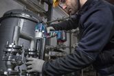 A person wearing safety equipment uses a cordless impact wrench on industrial piping.