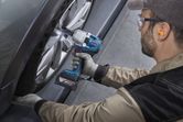 A person wearing safety equipment uses a cordless impact wrench on a car wheel.