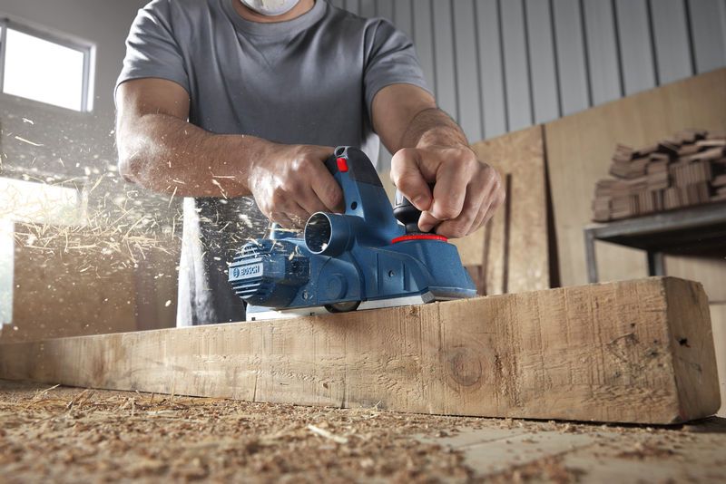A person wearing safety equipment planes a wooden beam, wood shavings flying.