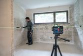 Person wearing safety equipment aligns wall features using a laser leveling tool in a tiled room.