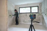 A worker wearing safety equipment aligns tiles using a laser leveling tool on a tripod.