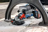 A person wearing safety equipment grinds concrete under a metal bench with a cordless tool.
