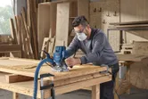 A person wearing safety equipment uses a router to shape wood in a workshop.